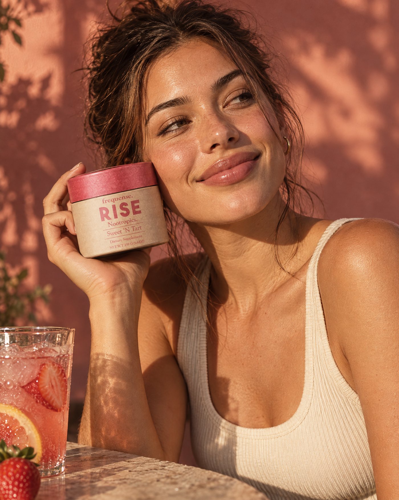 Woman in sunset light holding the RISE Sweet 'N Tart jar to her cheek, iced strawberry lemonade beside her.
