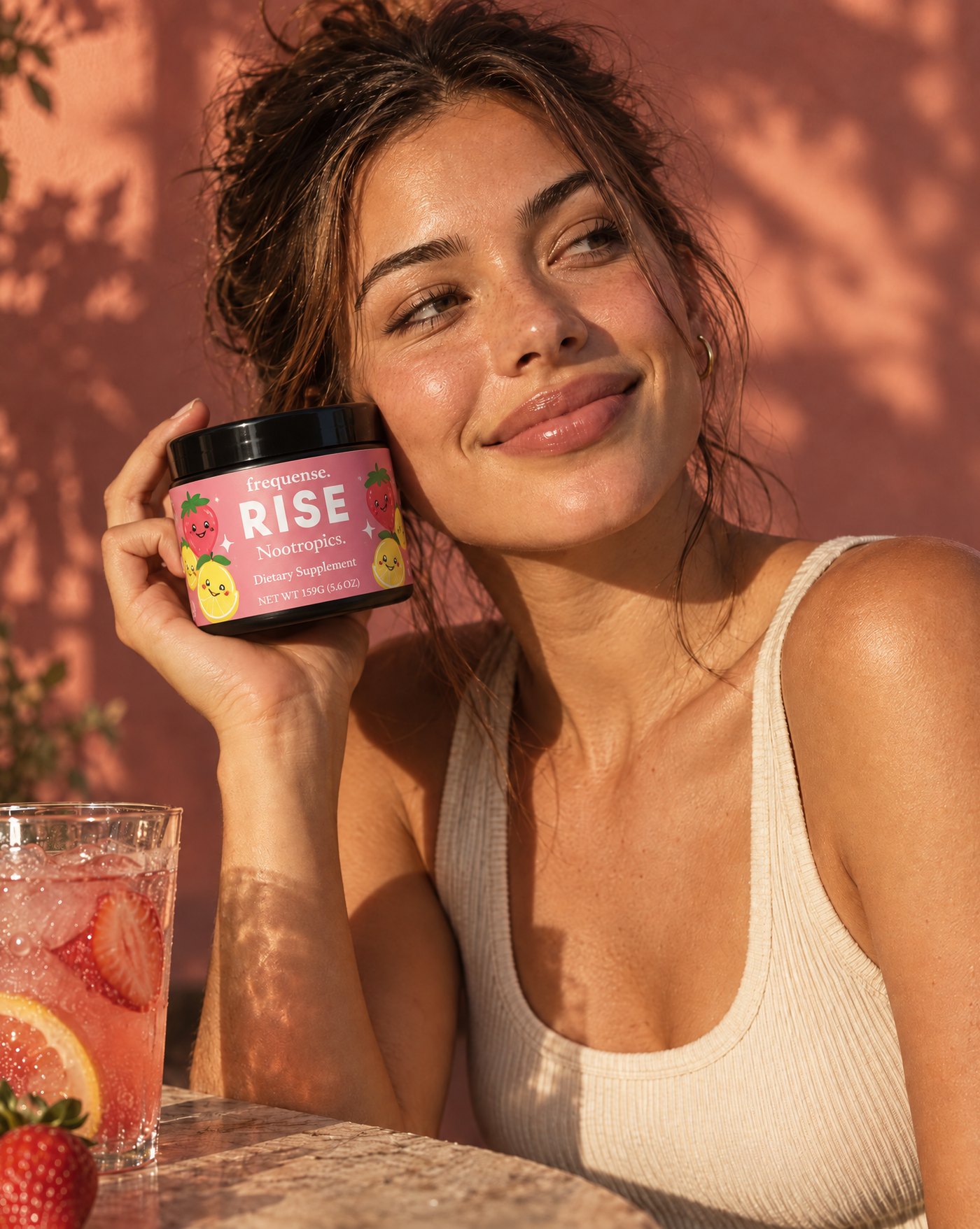 Woman in sunset light holding the RISE Sweet 'N Tart jar to her cheek, iced strawberry lemonade beside her.