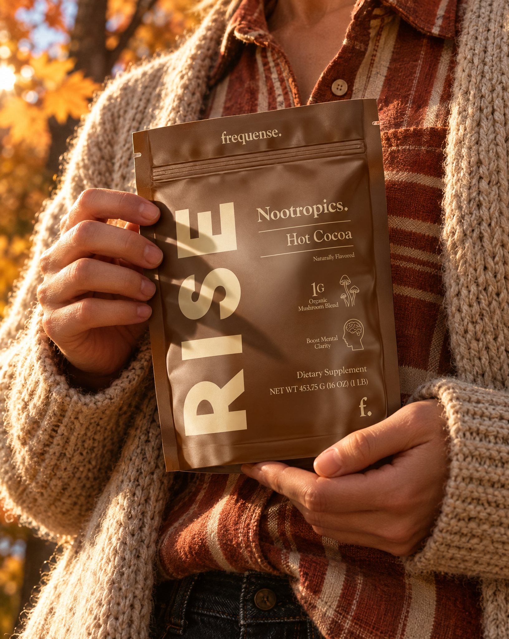 Hands in a cozy knit cardigan holding a RISE Hot Cocoa pouch under golden autumn leaves.