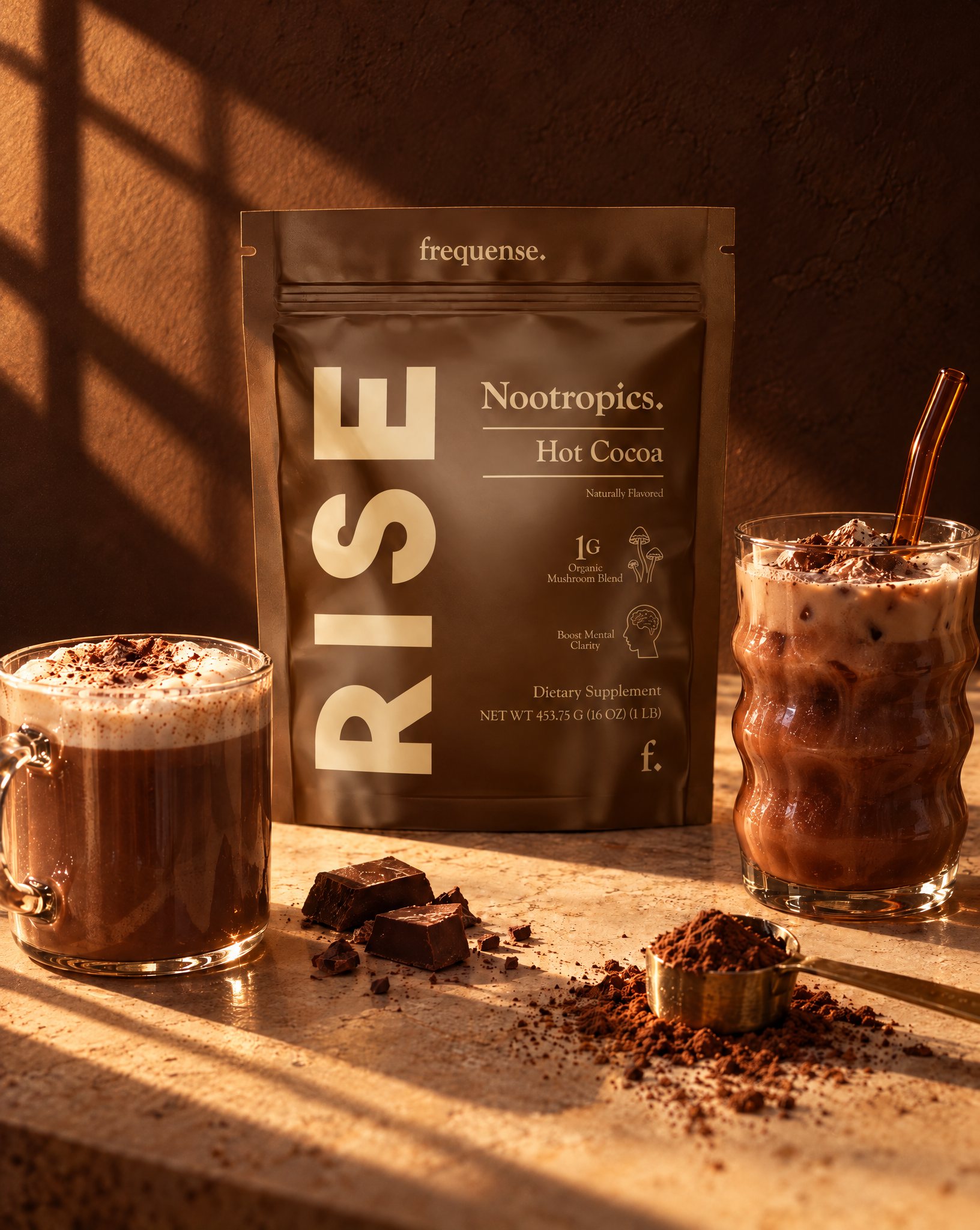 RISE Hot Cocoa pouch between a frothy hot mug and an iced cocoa with a glass straw, cocoa powder and chocolate on the counter.