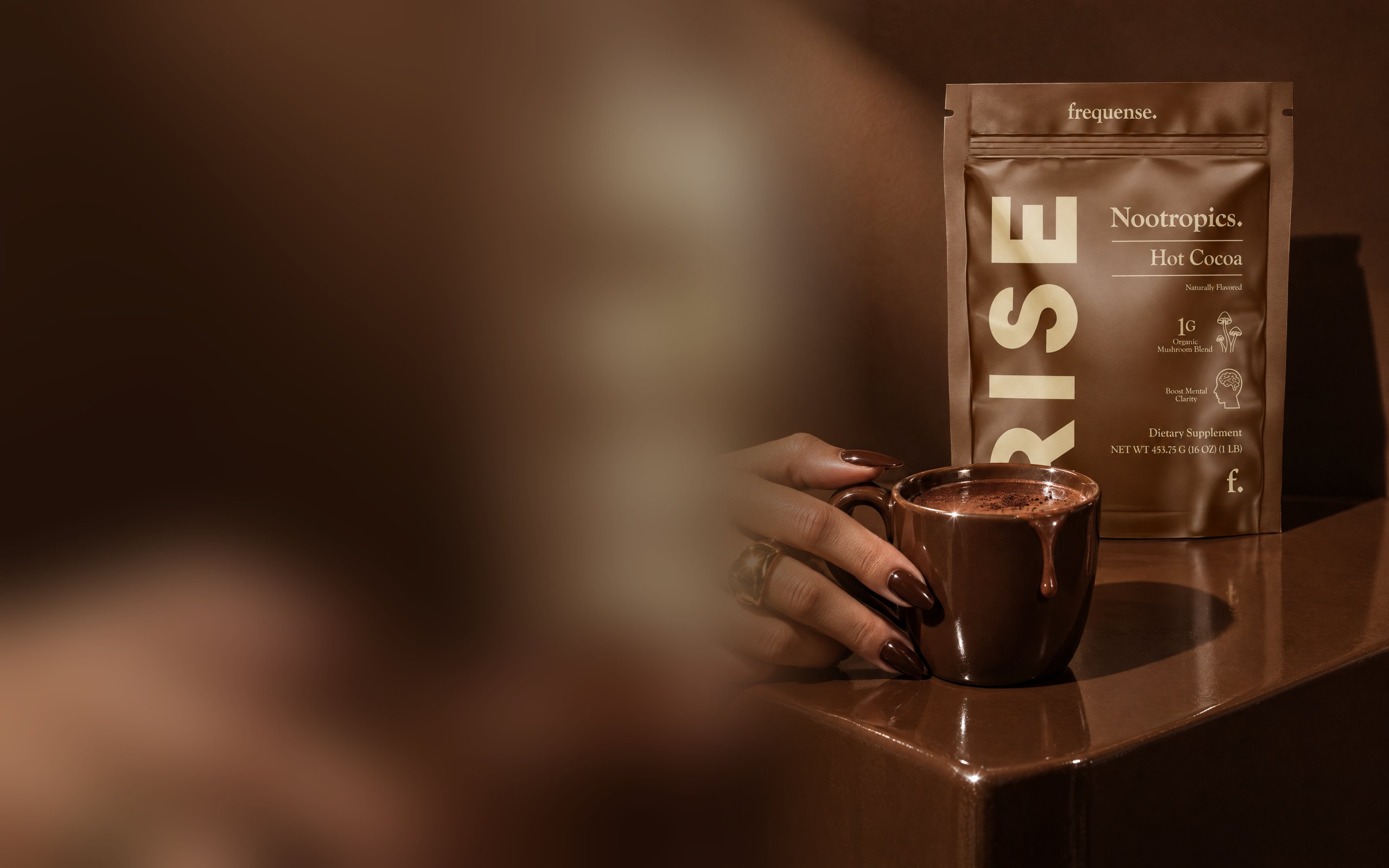 RISE Hot Cocoa pouch on a glossy espresso-brown ledge beside a hand cradling a dripping mug of rich hot cocoa.